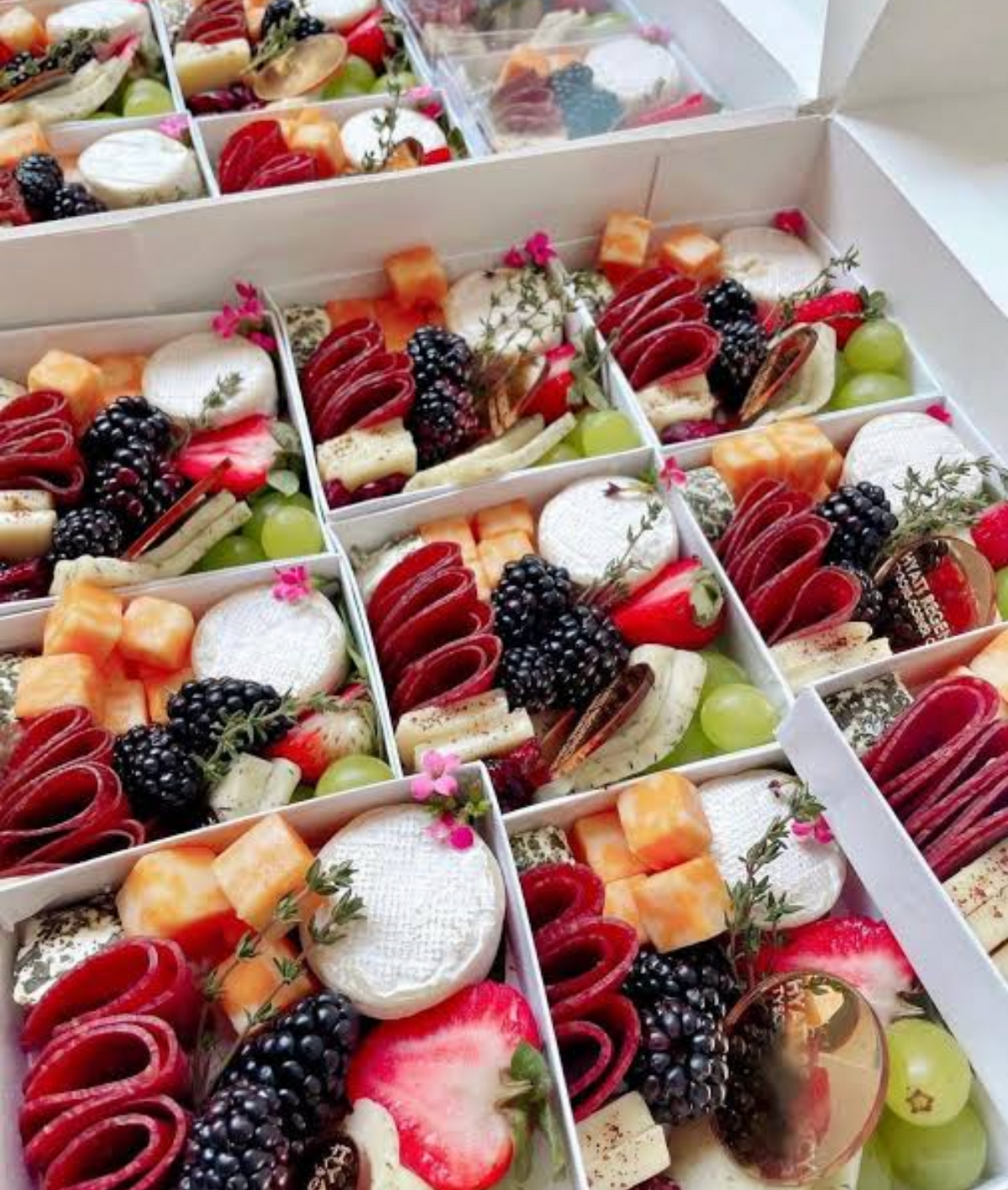 Mini charcuterie boxes filled with salami roses, brie, blackberries, strawberries, grapes, and edible flowers